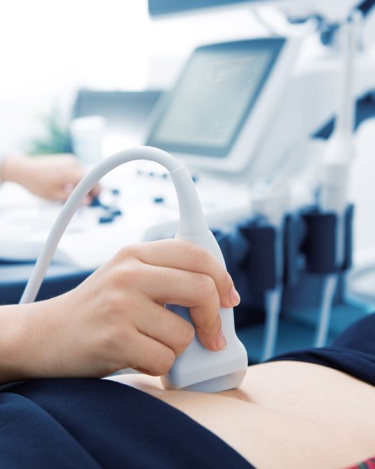 Głowica USG przykładana do brzucha Pacjenta, w tle zamazany sprzęt USG - ilustracja dotycząca USG pęcherza moczowego w Centrum Medycznym neoMedica w Poznaniu.