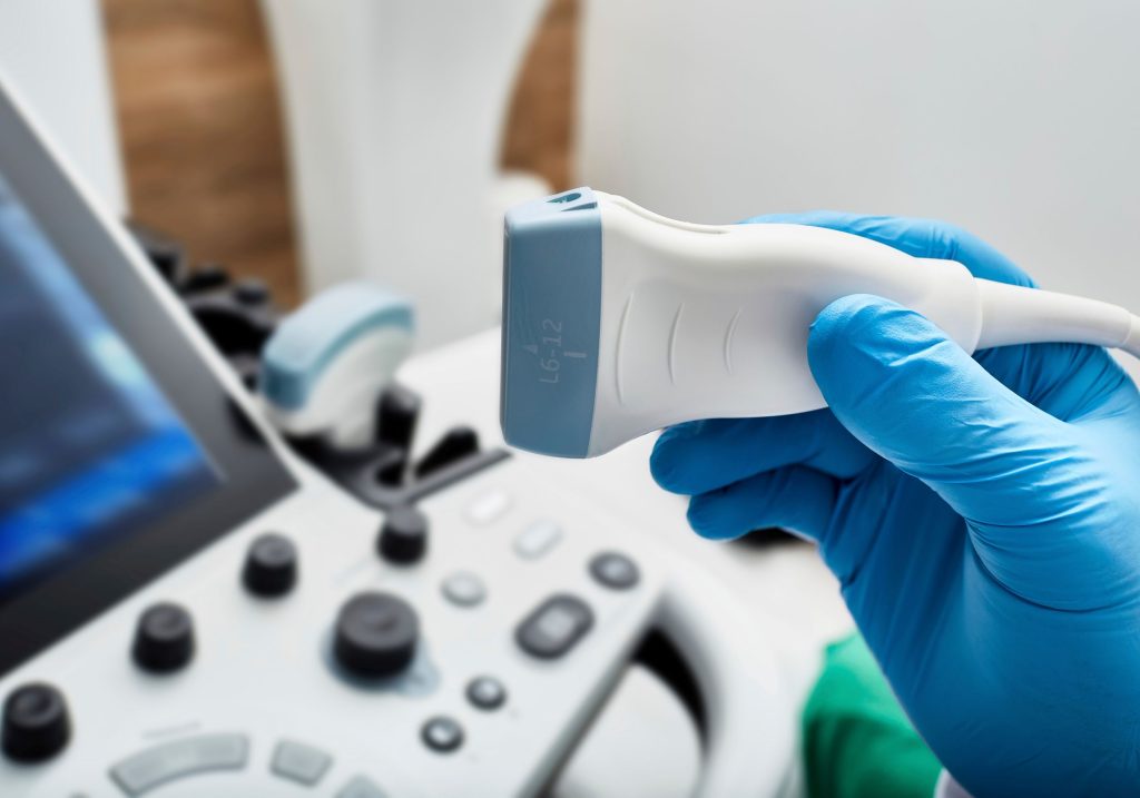 Ręka w rękawiczkach lekarskich trzyma głowicę USG, a w tle zamazany sprzet USG - ilustracja dotyczy USG jąder w Centrum Medycznym neoMedica w Poznaniu.