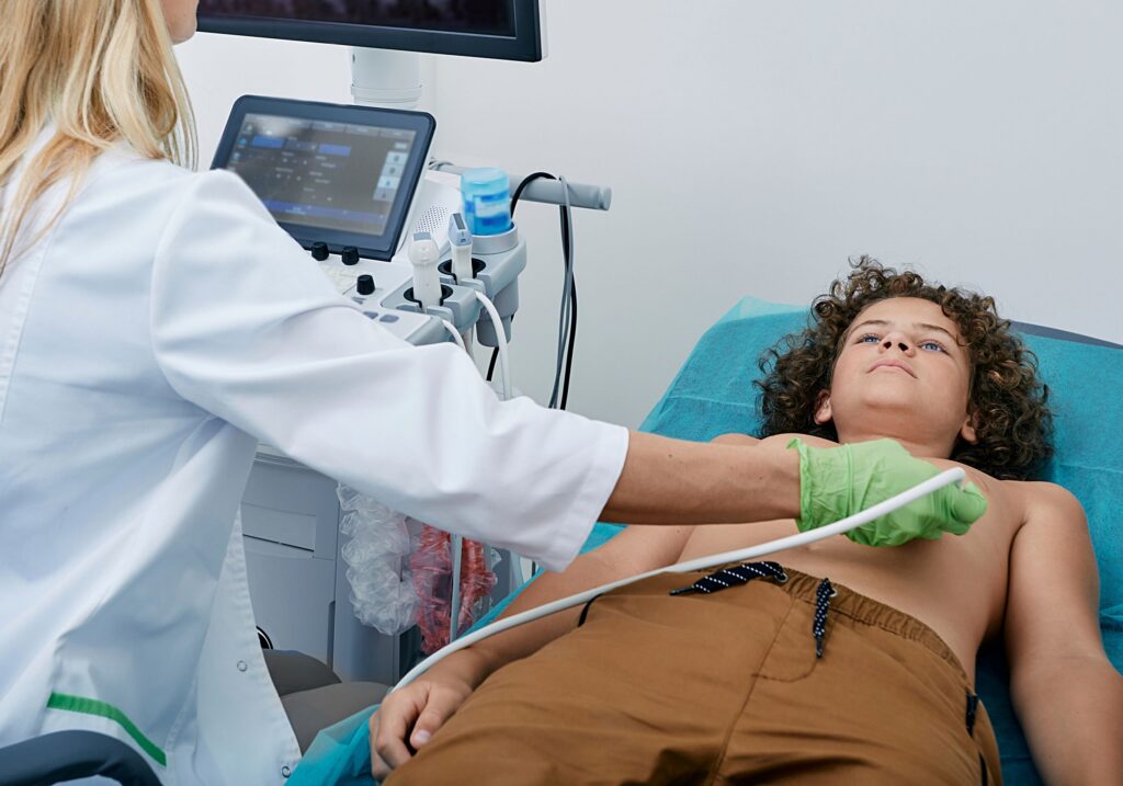 Leżący chłopiec z odsłoniętą klatką piersiową, a lekarz w rękawiczkach przykłada głowicę USG – ilustracja dotycząca badania echo serca dzieci w Centrum Medycznym neoMedica w Poznaniu.